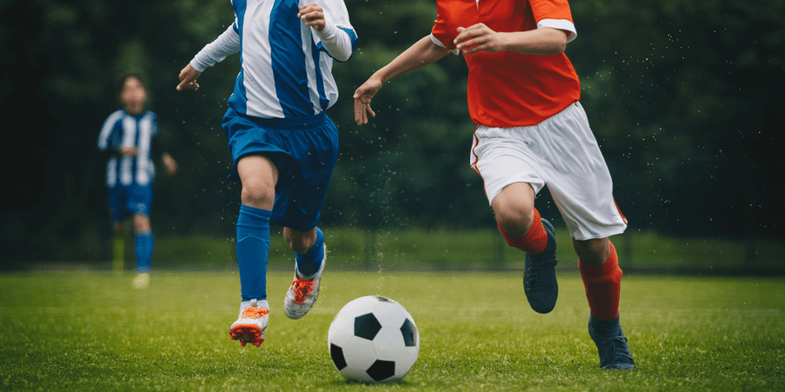 Two soccer players in blue and red jerseys running after a ball on a grassy field.