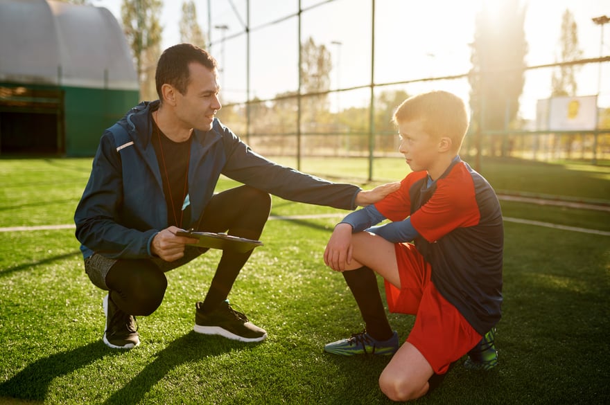 Youth soccer coach kneeling on the field, placing a supportive hand on a young player's shoulder during a one-on-one conversation before a game.