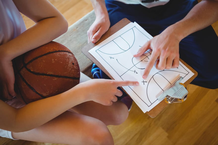 A coach and a benched athlete sitting together reviewing a basketball strategy on a clipboard, focusing on helping benched athlete overcome a performance slump.