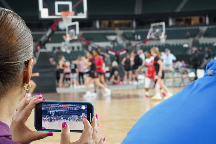 Parent recording a youth basketball game on a smartphone from courtside, capturing player stats and action on the arena floor.