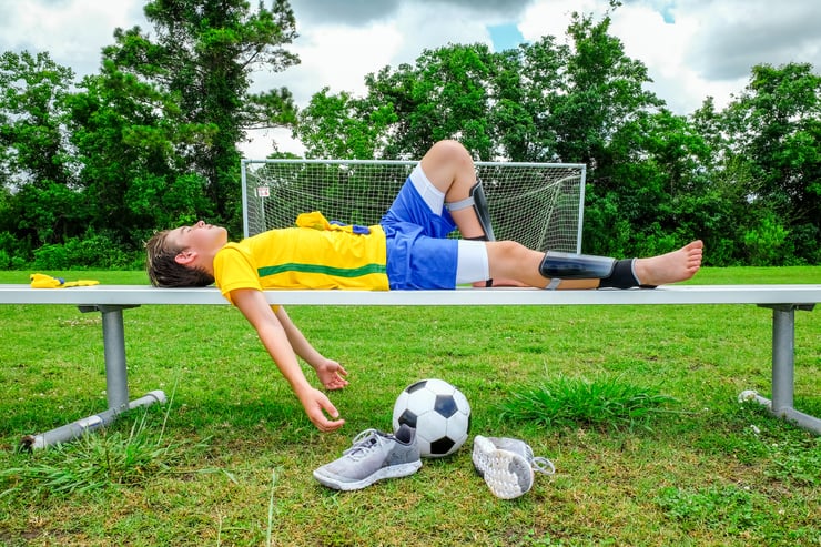 Two young athletes in yellow and blue uniforms lying exhausted on park bench after off-season training workout with soccer ball and shoes on ground